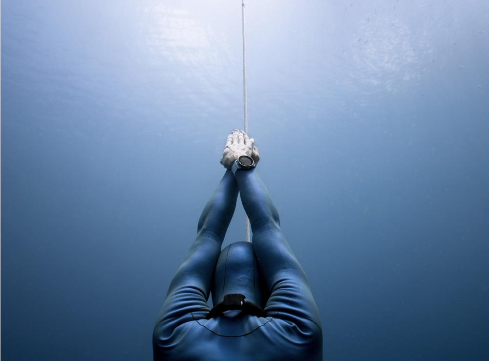 A freediver descends head-first along the line into deep blue water, hands together above the head