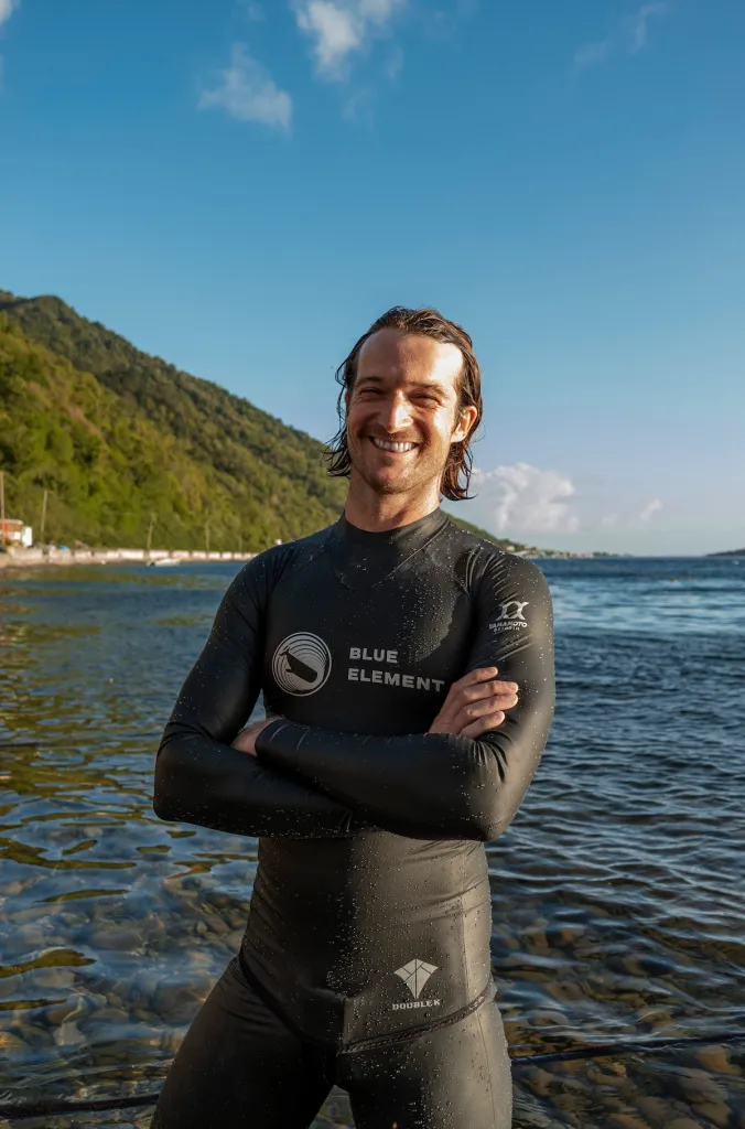 Harry McCahill smiling in his black Double K wetsuit with the Blue Element logo, Soufrière Bay behind